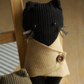 A black ribbed knit cat plush toy wearing a beige polka-dot shirt with a brown button, posed on a wooden chair next to another olive green plush toy.